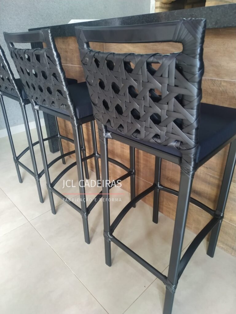 A imagem mostra três banquetas de bar alinhadas, com estrutura de alumínio pintado de preto e assentos revestidos com tecido escuro, proporcionando um contraste elegante. O encosto das banquetas é feito de fibra sintética entrelaçada, com acabamento sofisticado e moderno. Ao fundo, é possível ver uma bancada de cozinha de granito escuro, complementando o ambiente com estilo e funcionalidade. O piso claro e as paredes texturizadas realçam a simplicidade e sofisticação do espaço. O logo da JCL Cadeiras, especializada na fabricação e reforma de móveis, é visível, destacando a qualidade do produto.